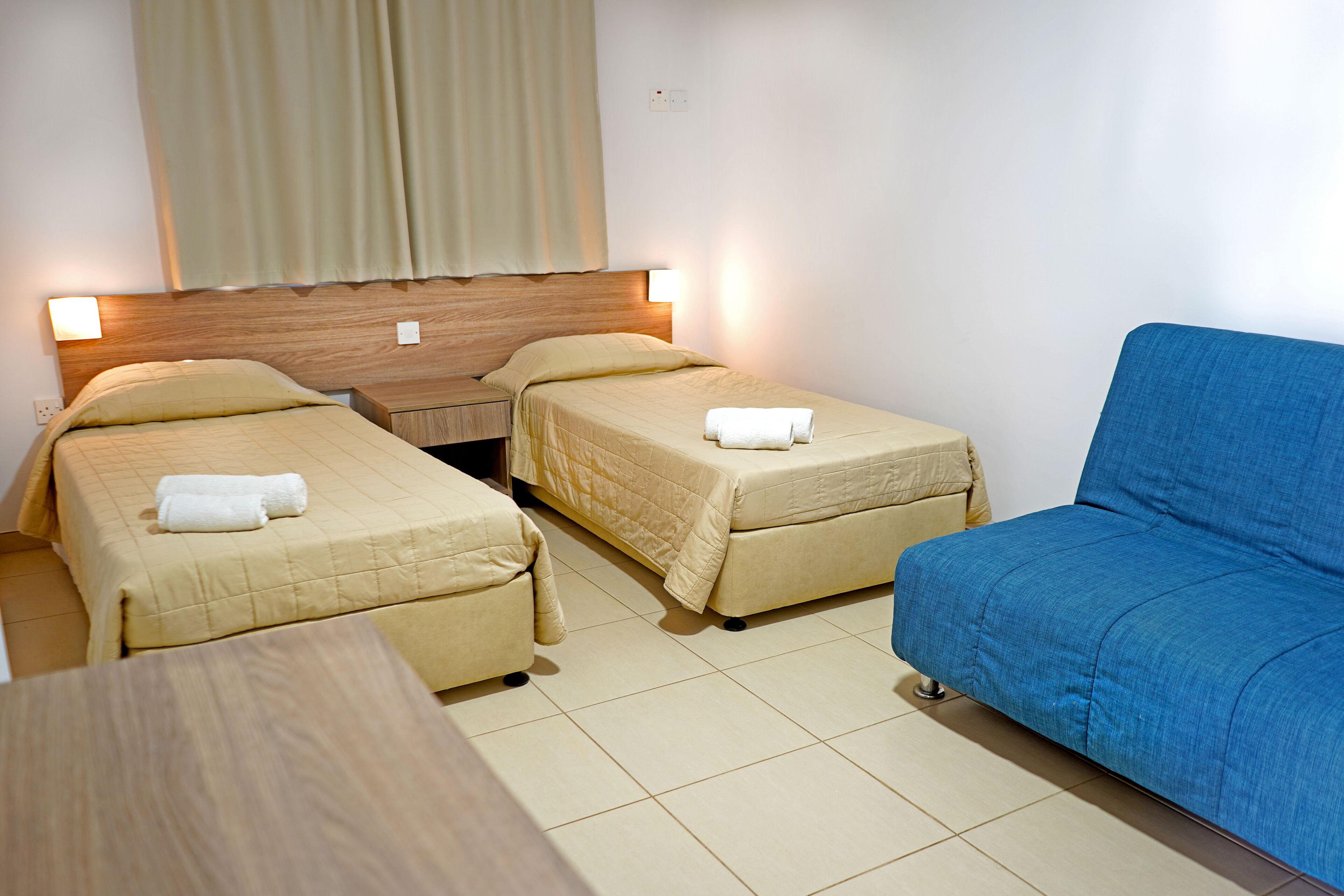 Habitación de hotel con dos camas individuales, un sofá, una mesa de madera y una ventana con cortinas.