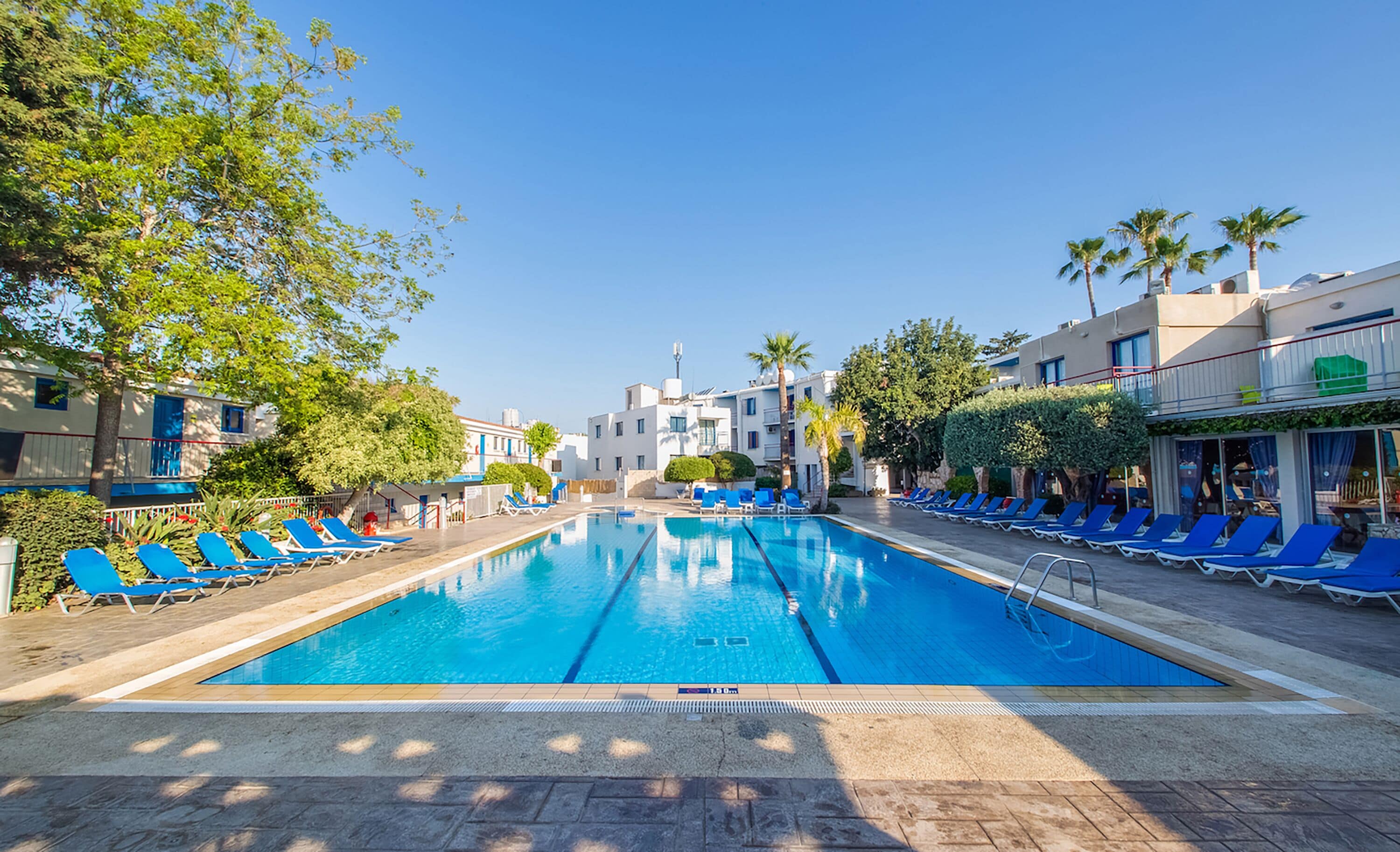 A swimming pool with blue lounge chairs on both sides, surrounded by buildings with balconies and palm trees.