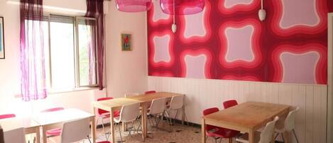 Una habitación con mesas y sillas, un ventanal con cortinas, y una pared con un diseño geométrico.