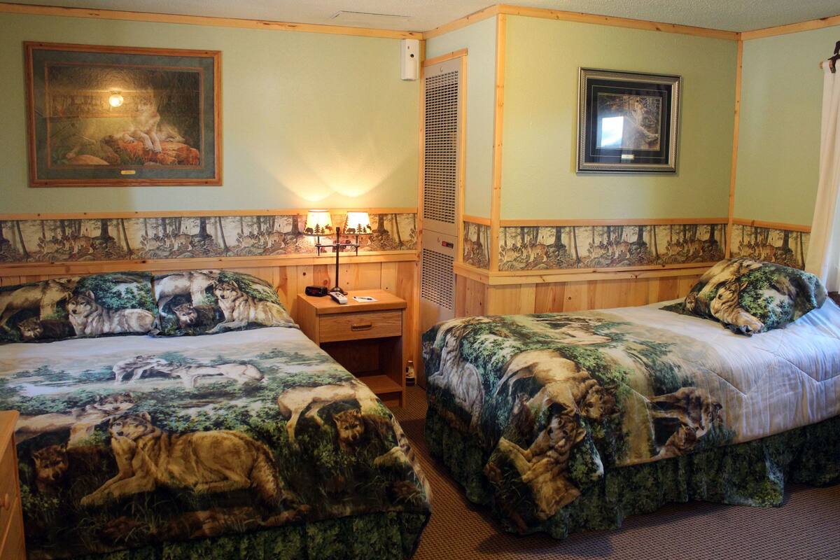 A room with two beds, each with a forest-themed bedspread. There is a nightstand with a lamp and a telephone. The walls have a painted border depicting wolves and deer.