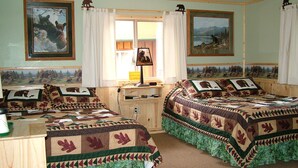A room with two beds, each with a quilted bedspread featuring animal prints. There are two framed pictures on the walls, a window with curtains, and a desk with a lamp. - Glacier Haven Inn (Essex)