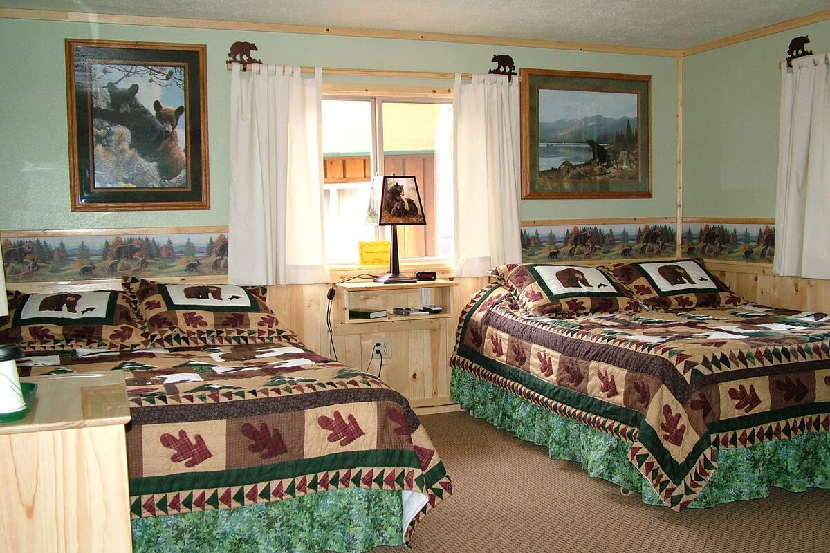A room with two beds, each with a quilted bedspread featuring animal prints. There are two framed pictures on the walls, a window with curtains, and a desk with a lamp.