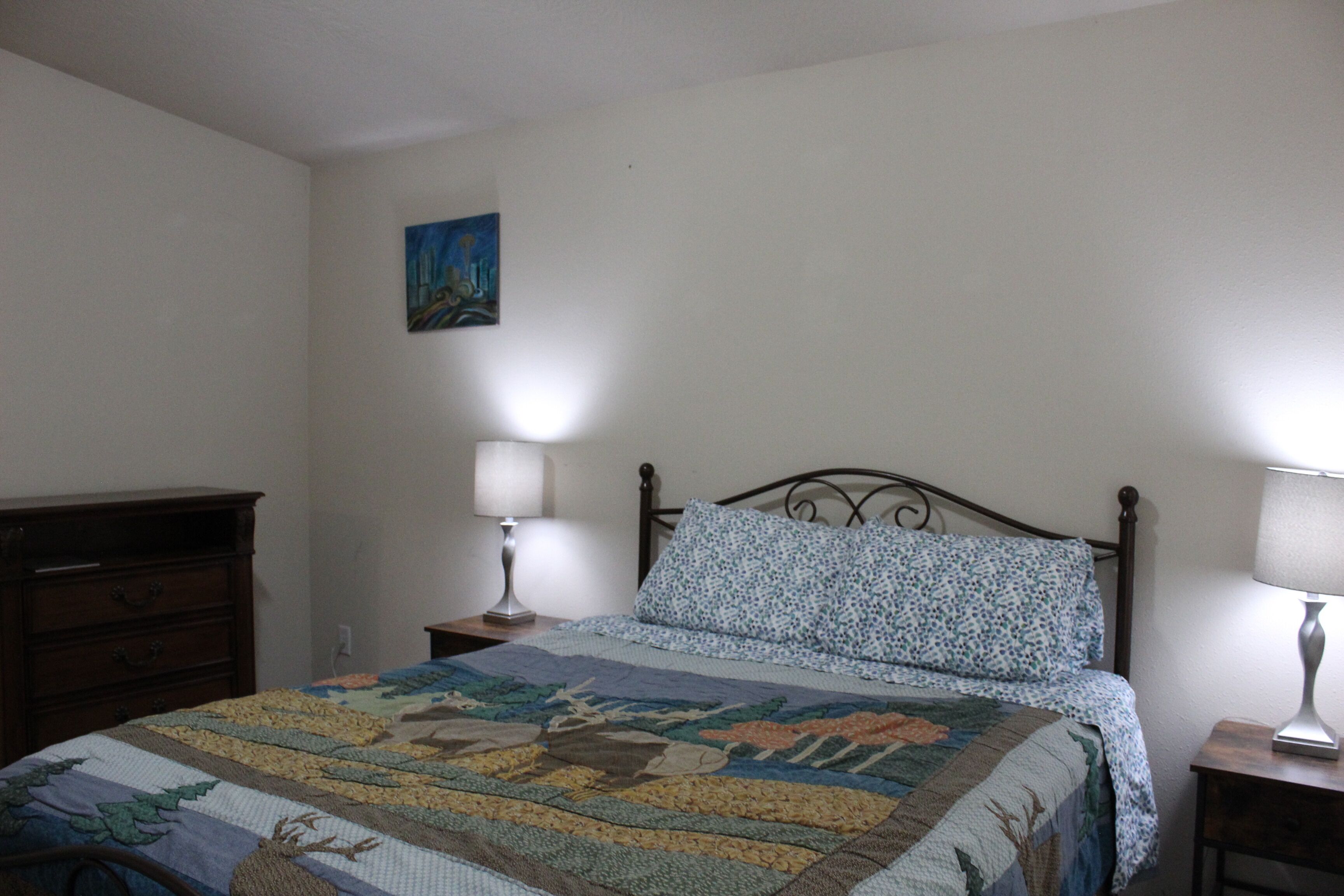 A bedroom with a bed, a dresser, two bedside lamps, and a painting on the wall.