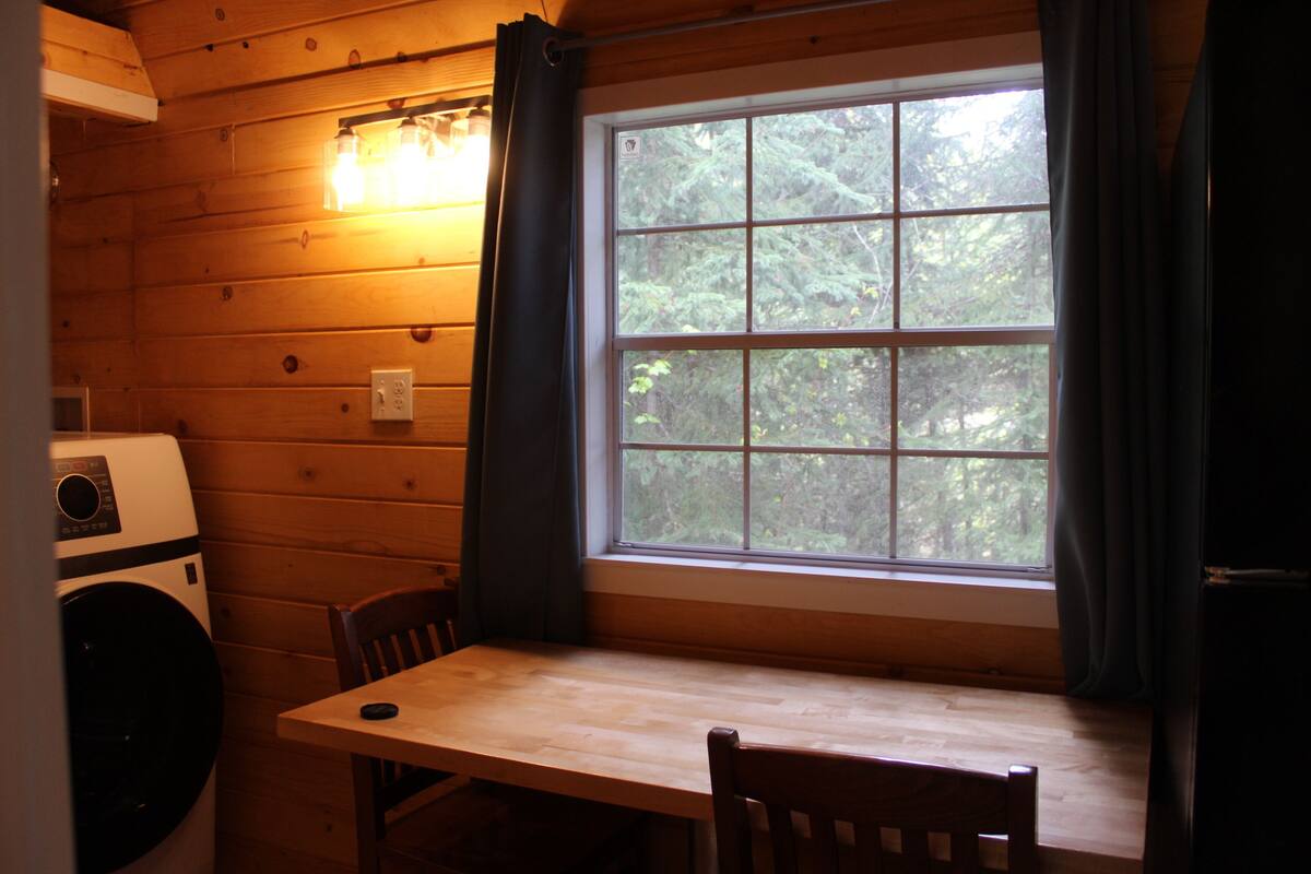 A room with a wooden interior, a window with curtains, a table, and a washing machine.