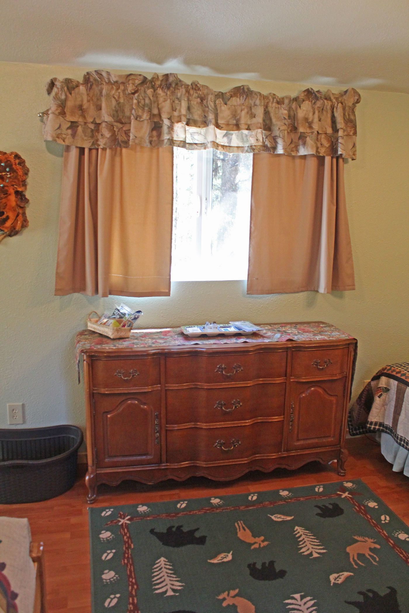 A room with a wooden dresser, a window with curtains, a patterned rug, and a small basket on the dresser.