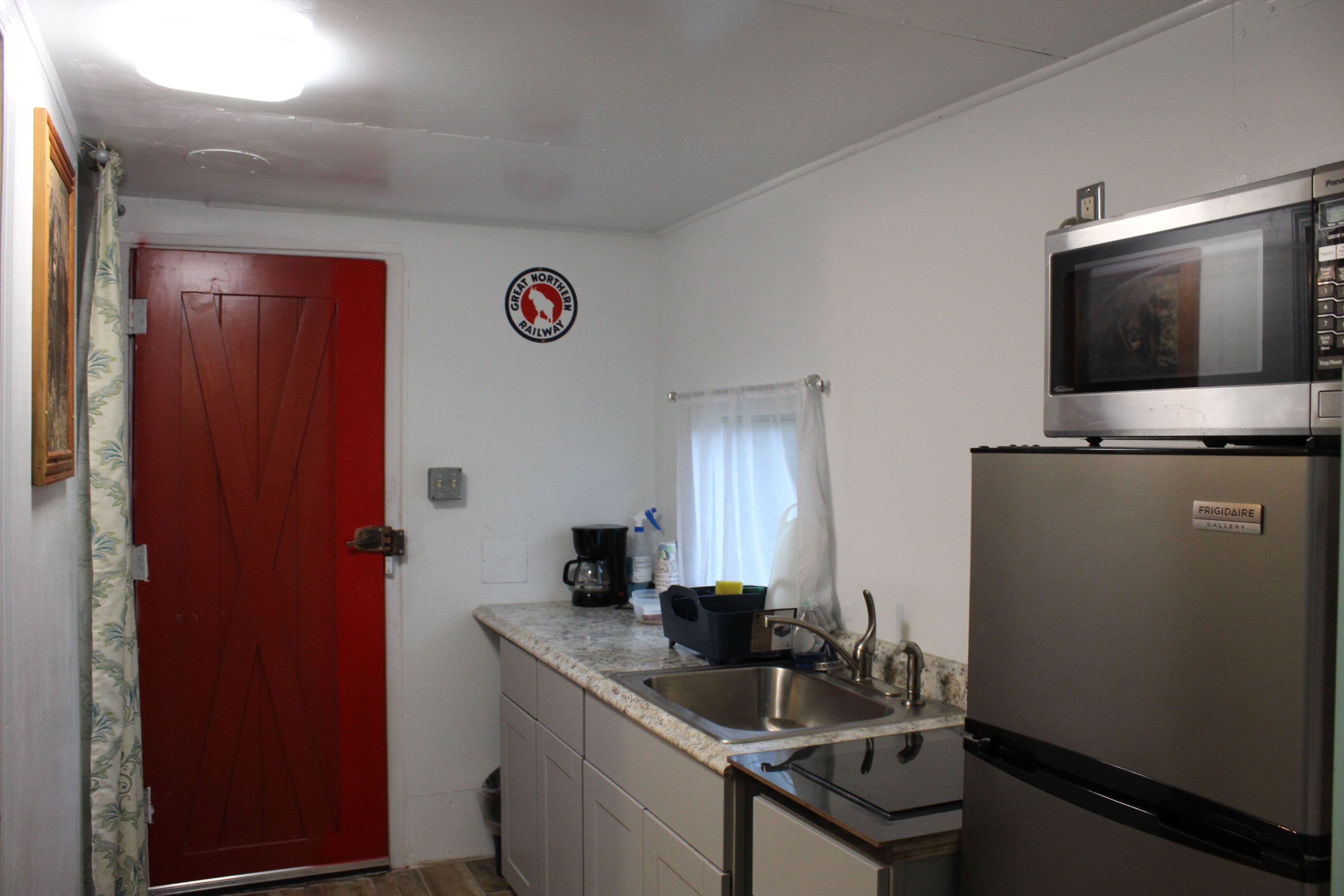 A compact kitchen with a red door, a stainless steel refrigerator, a microwave, and a coffee maker.