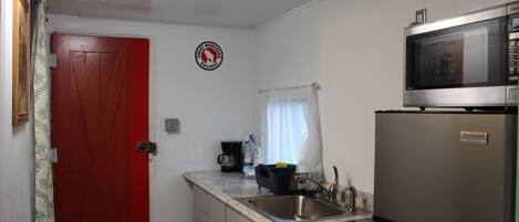 A compact kitchen with a red door, a stainless steel refrigerator, a microwave, and a coffee maker.
