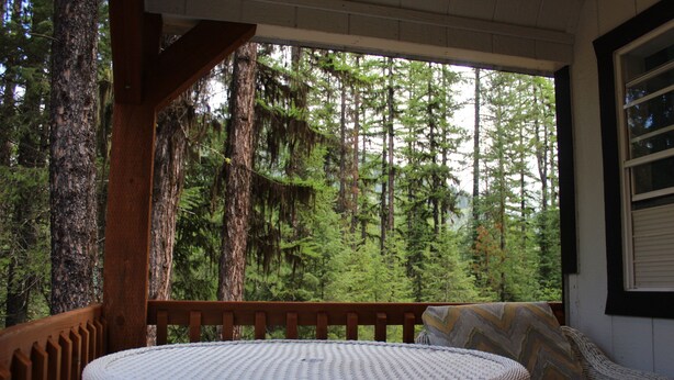 A wooden deck with a round table and a cushioned bench, overlooking a dense forest. - Glacier Haven Inn (Essex)
