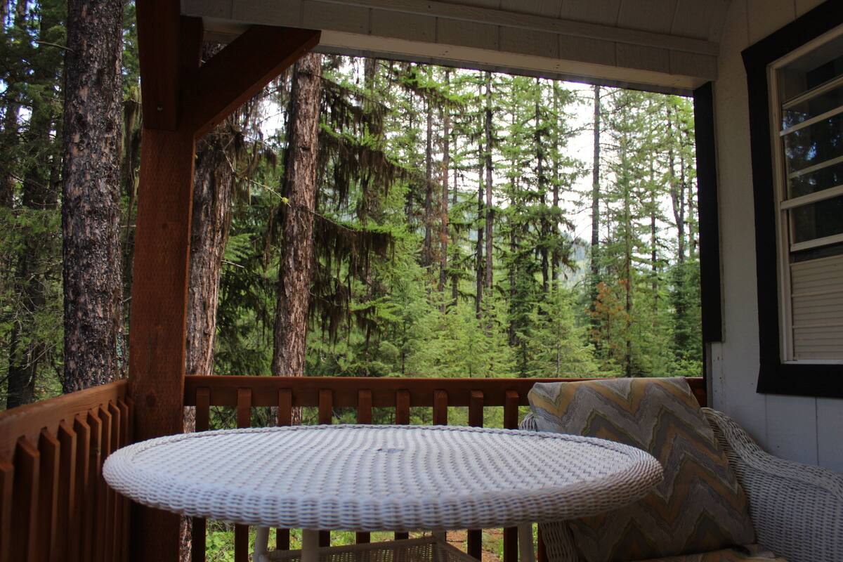 A wooden deck with a round table and a cushioned bench, overlooking a dense forest.