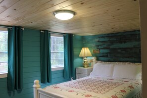 A bedroom with teal walls, a wooden ceiling, a bed with a floral bedspread, a nightstand with a lamp, and a window with curtains.