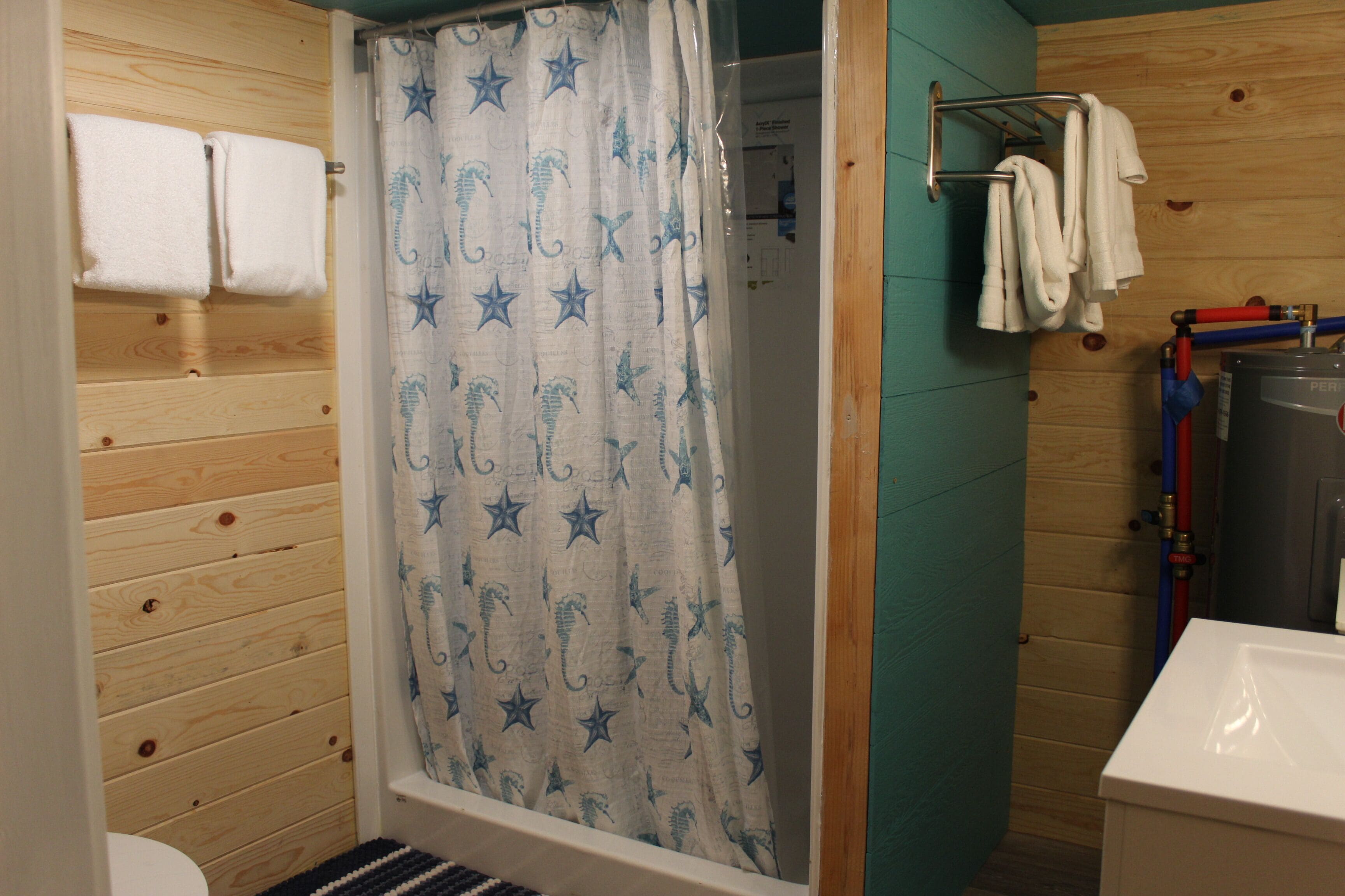 Un baño con paneles de madera, cortina de ducha con motivos de estrellas, toallero con toallas y un calentador de agua visible.