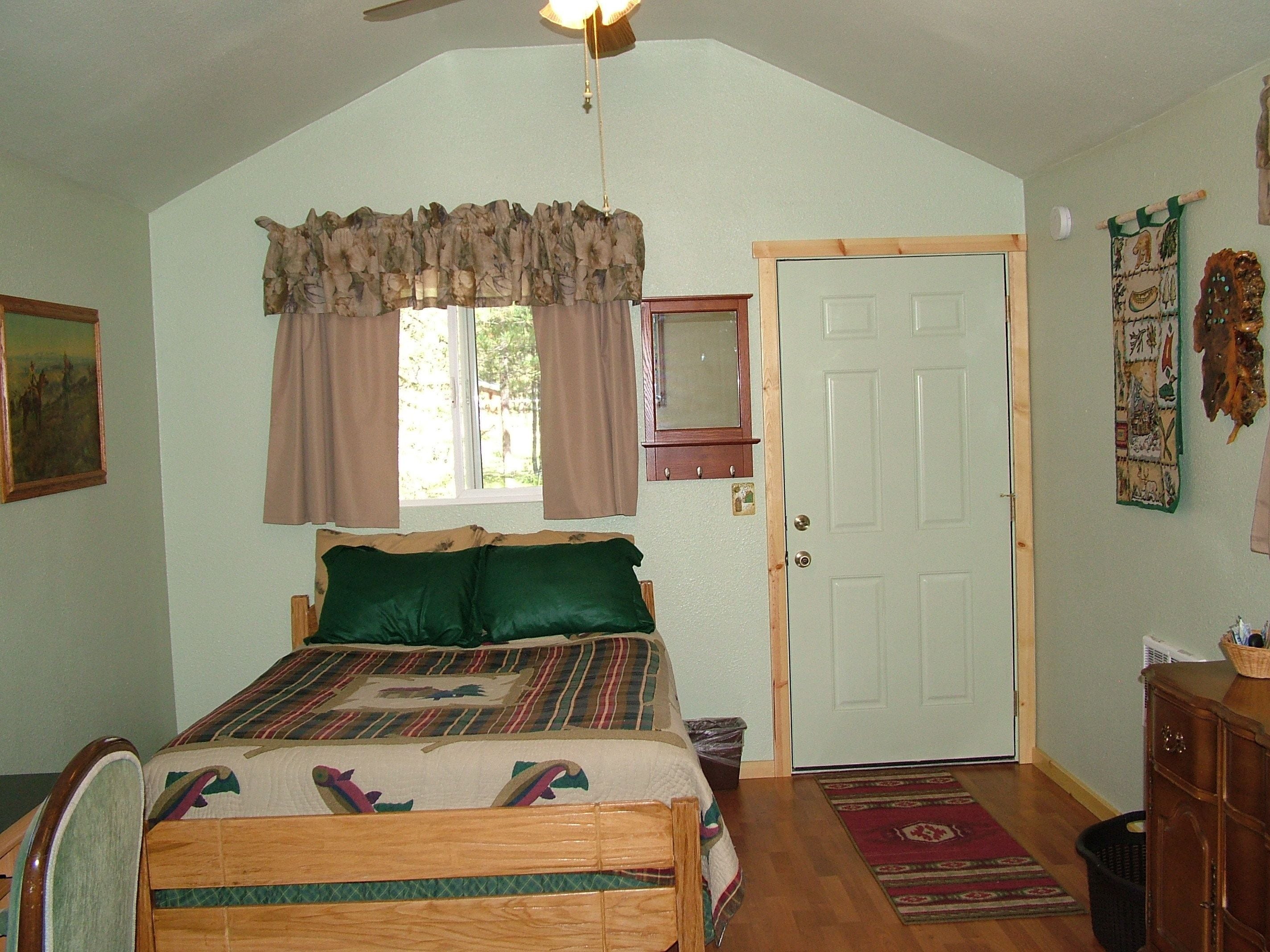 Un dormitorio con cama, ventana con cortinas, puerta, ventilador de techo y espejo montado en la pared, además de una alfombra.