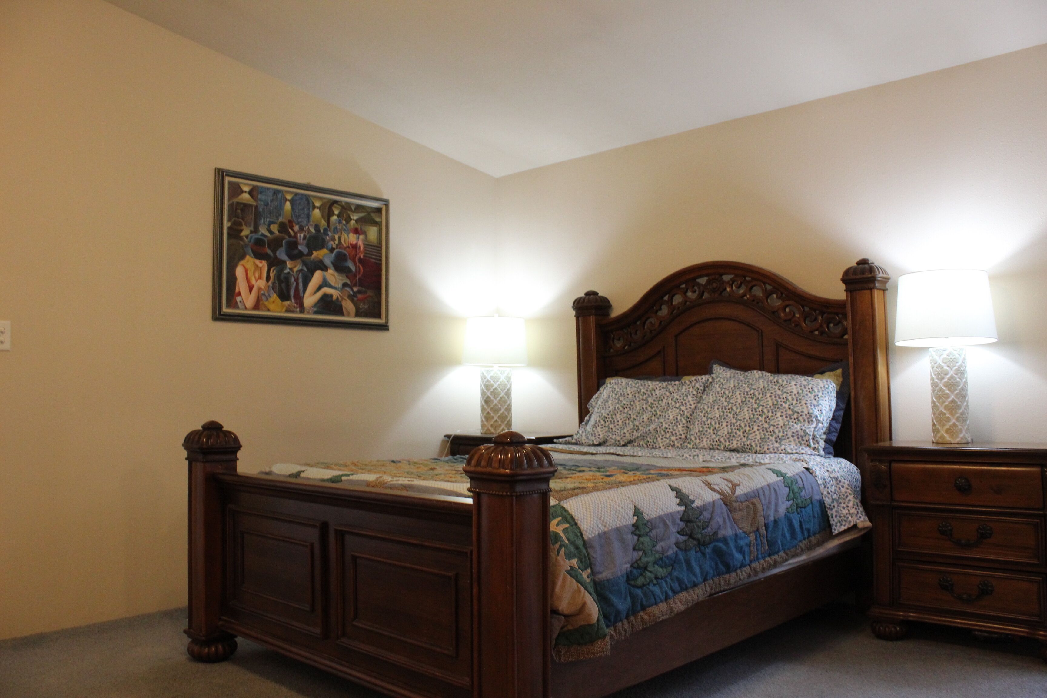 A bedroom with a wooden bed, two bedside tables with lamps, a wall-mounted painting, and a carpeted floor.