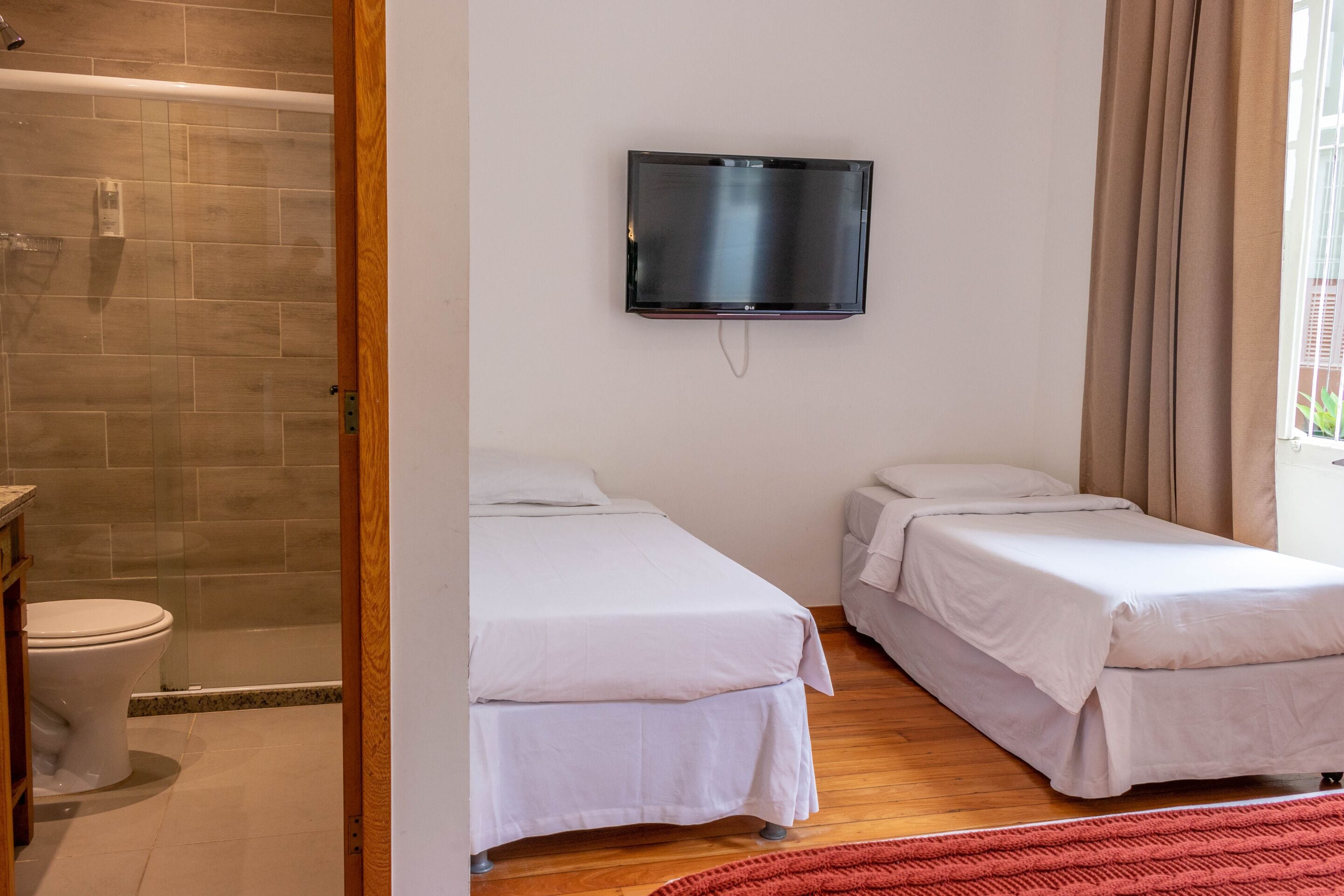 Habitación de hotel con dos camas, televisor montado en la pared, zona de ducha y ventana con cortinas.
