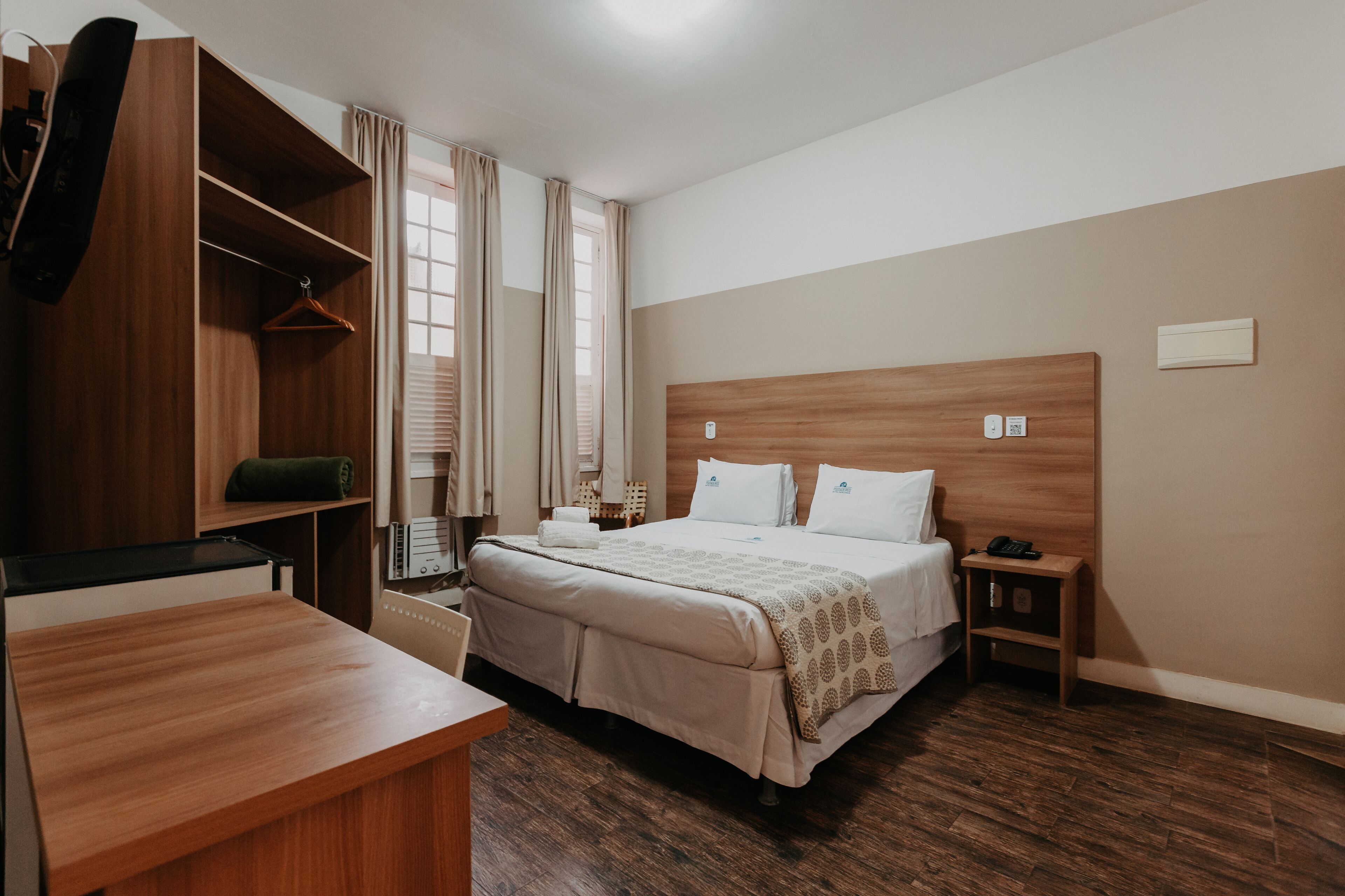 A hotel room with a wooden headboard, a bed with a patterned blanket, a wooden desk, a wardrobe, and a bedside table with a phone.