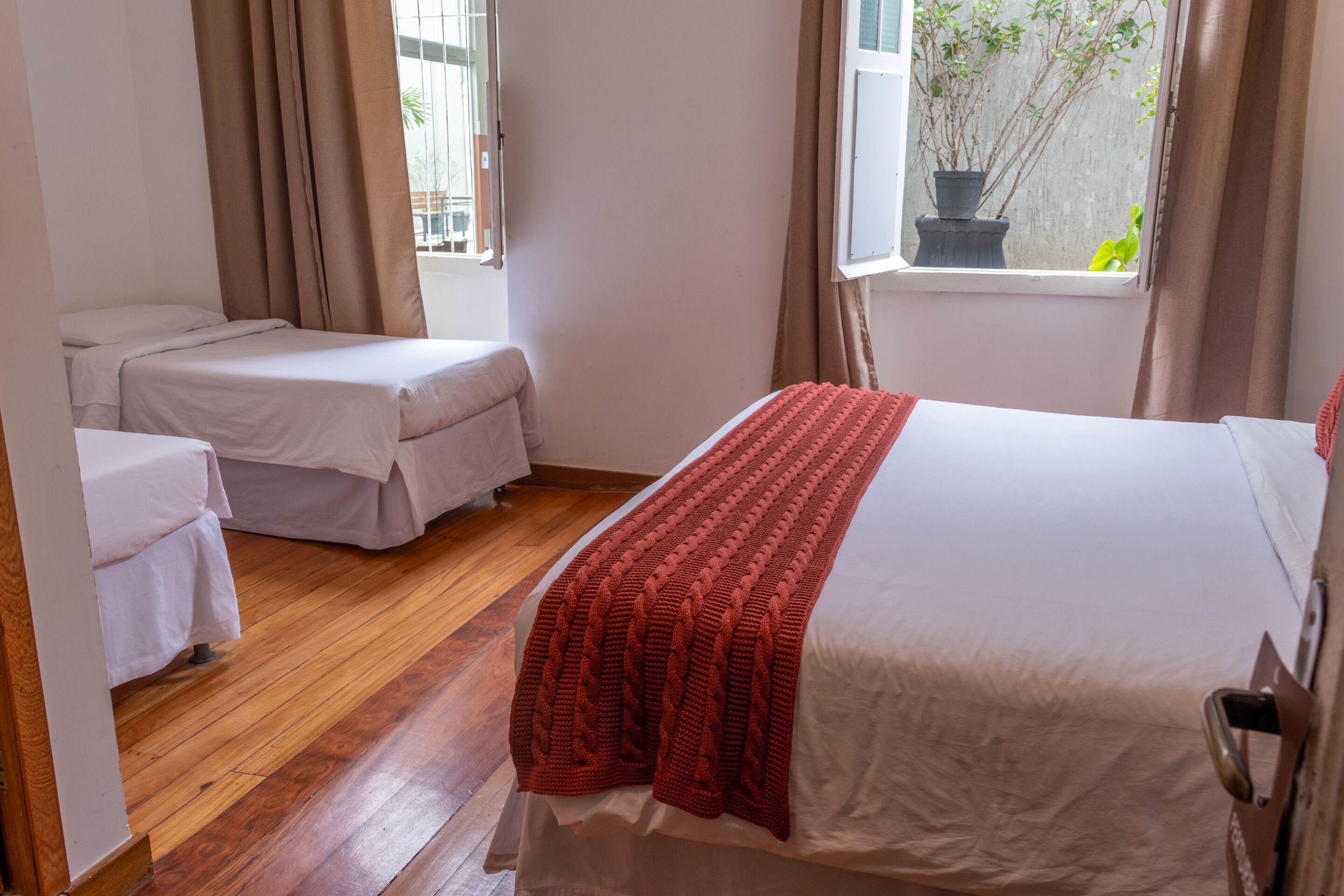 A hotel room with a large bed, a smaller bed, wooden flooring, and a window with a view of greenery.