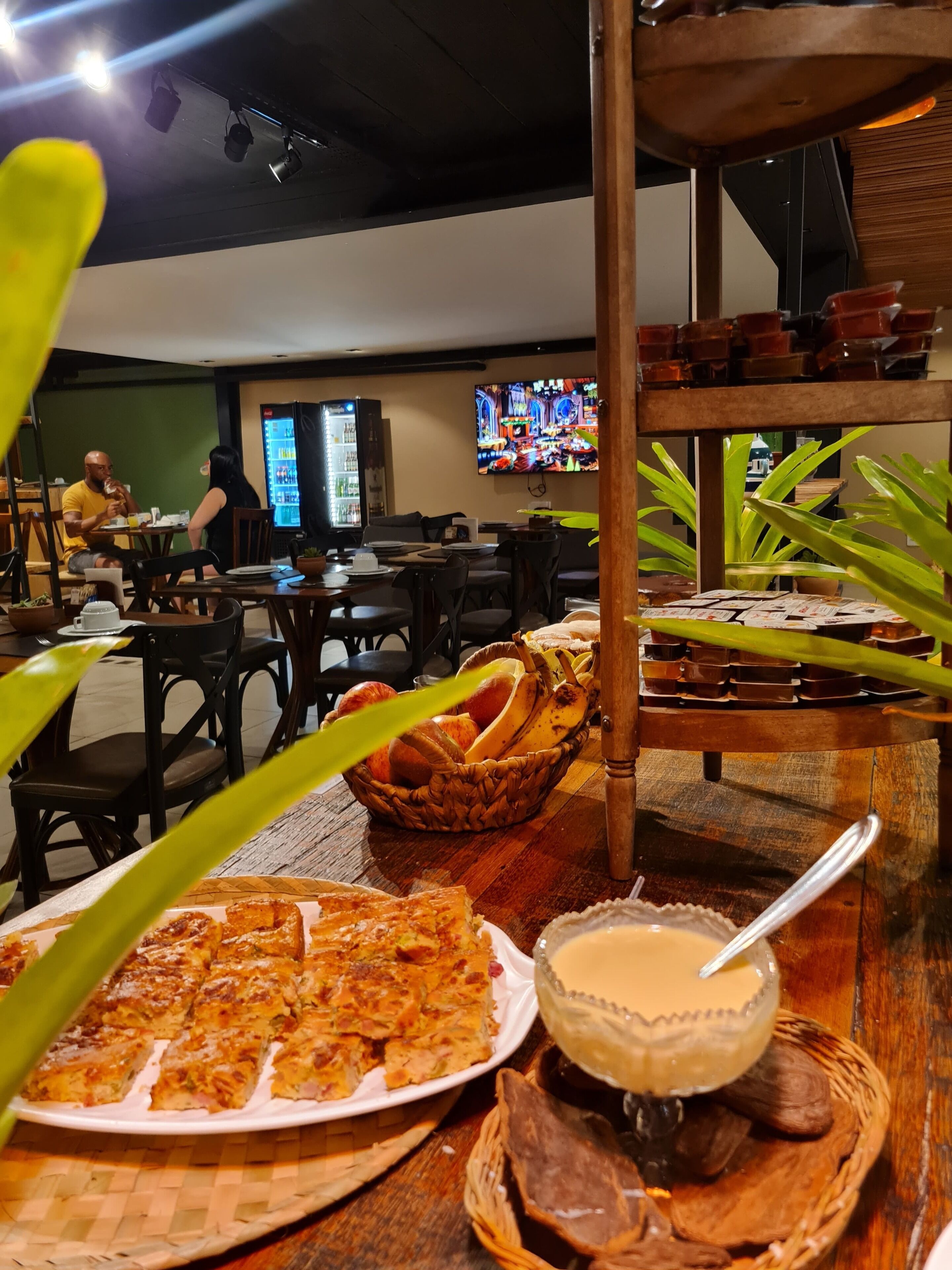 A restaurant with tables, chairs, a television screen, and a variety of food items including a pizza and a drink.