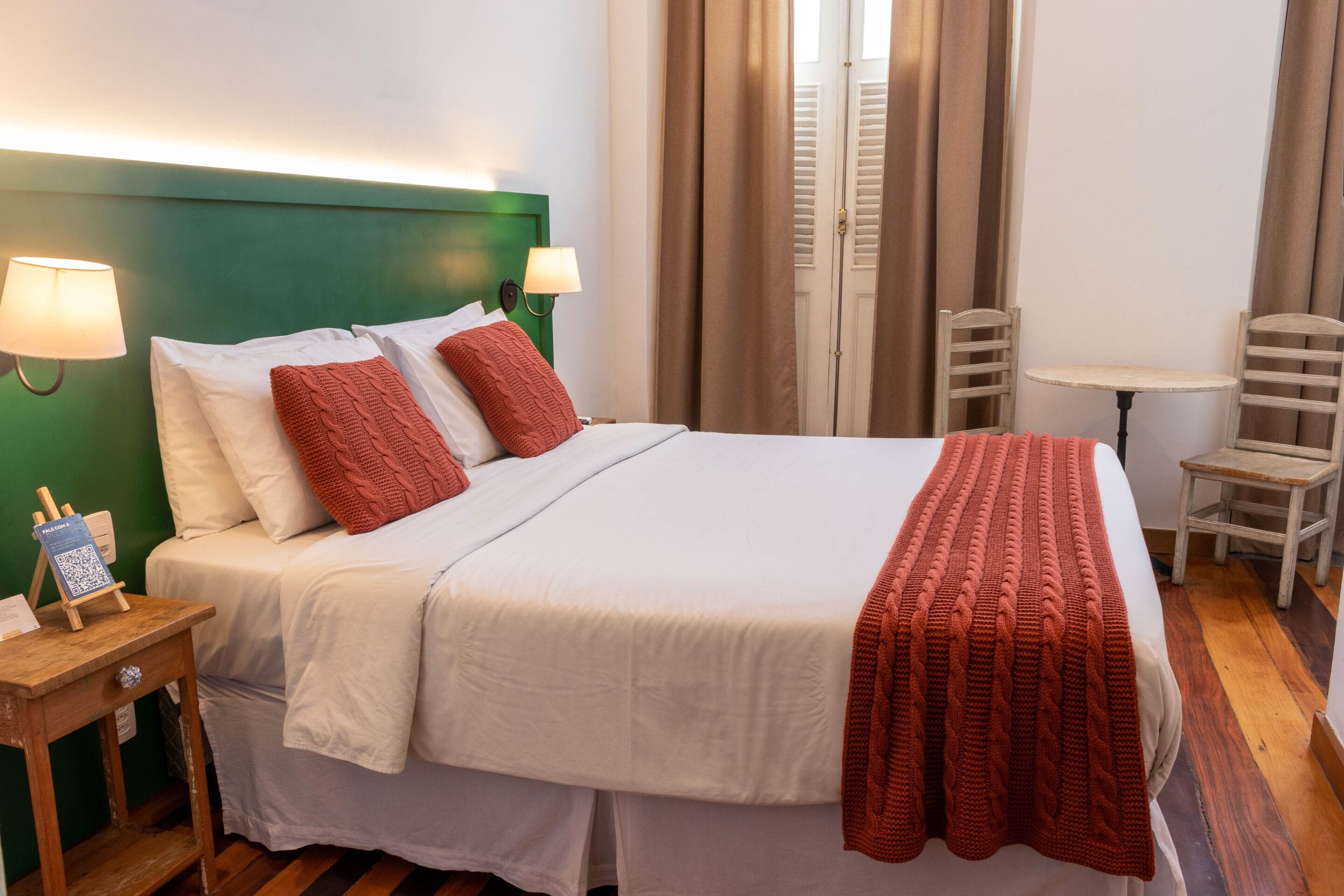 A neatly made bed with white linens and red accents, a bedside table with a lamp, and a small table with chairs in a room with wooden flooring and green walls.