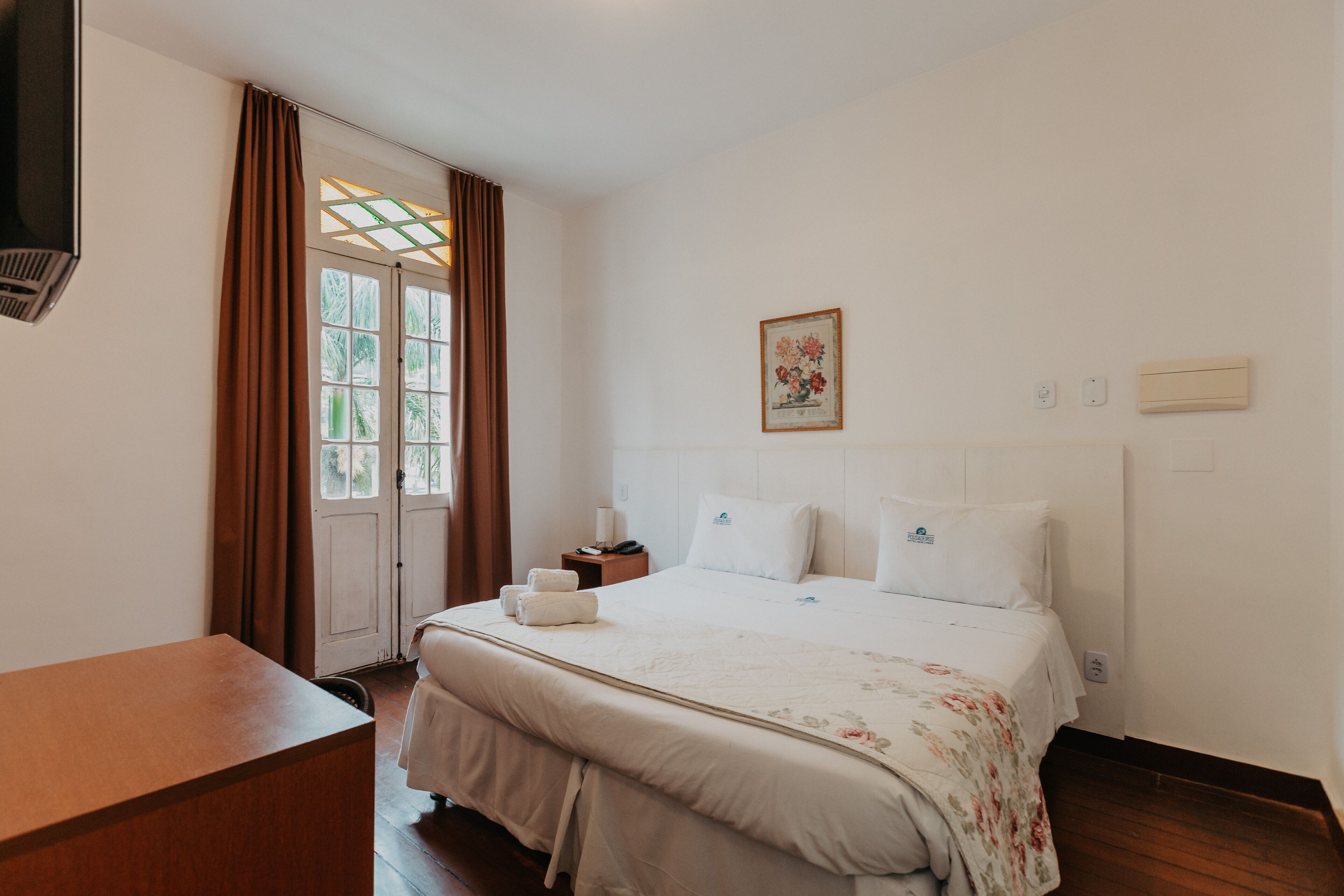A hotel room with a bed, a bedside table, a desk, a window with curtains, and a wall-mounted air conditioner.