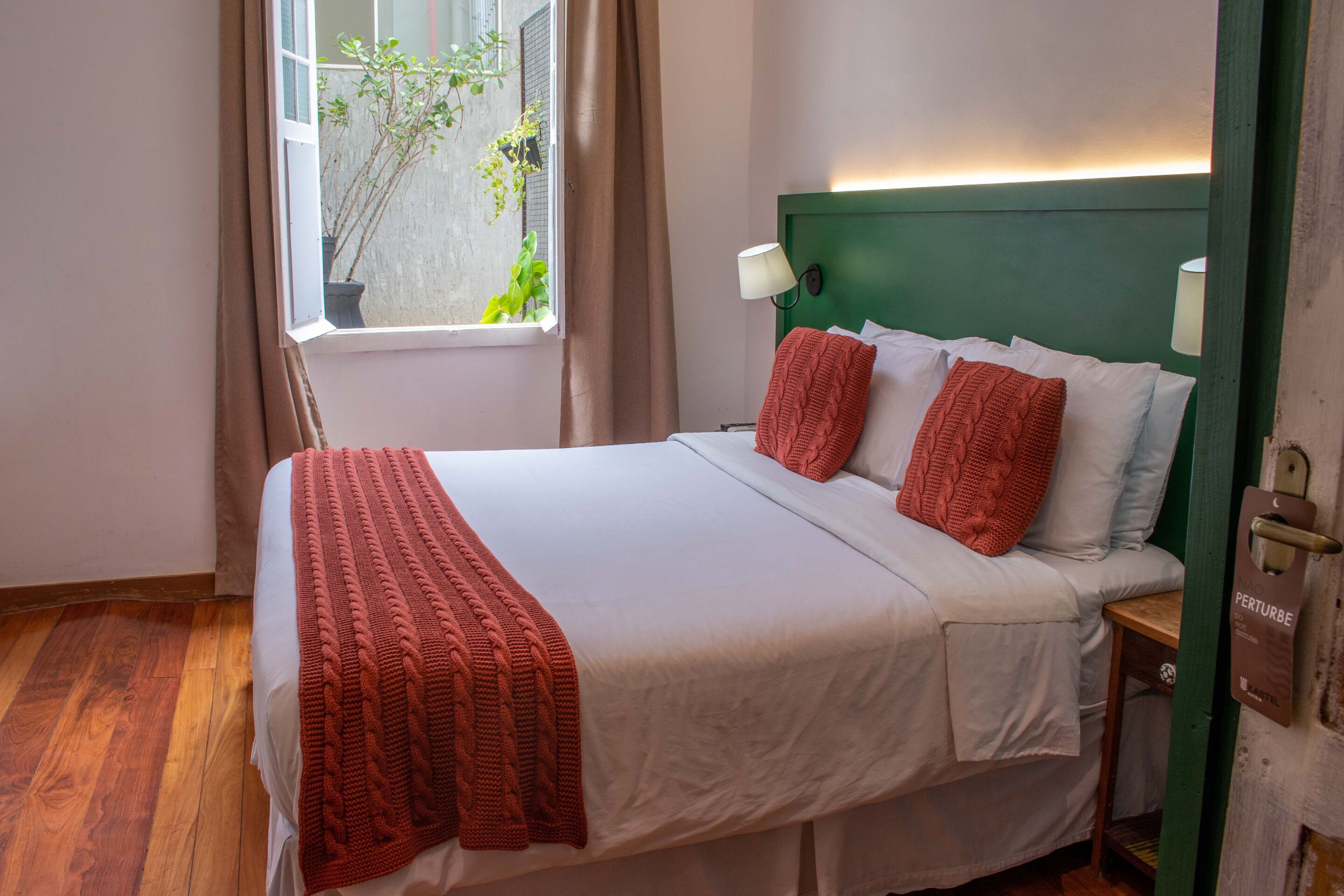 Een netjes opgemaakt bed met witte lakens en rode accenten, een bedlampje, en uitzicht op groen door een raam.