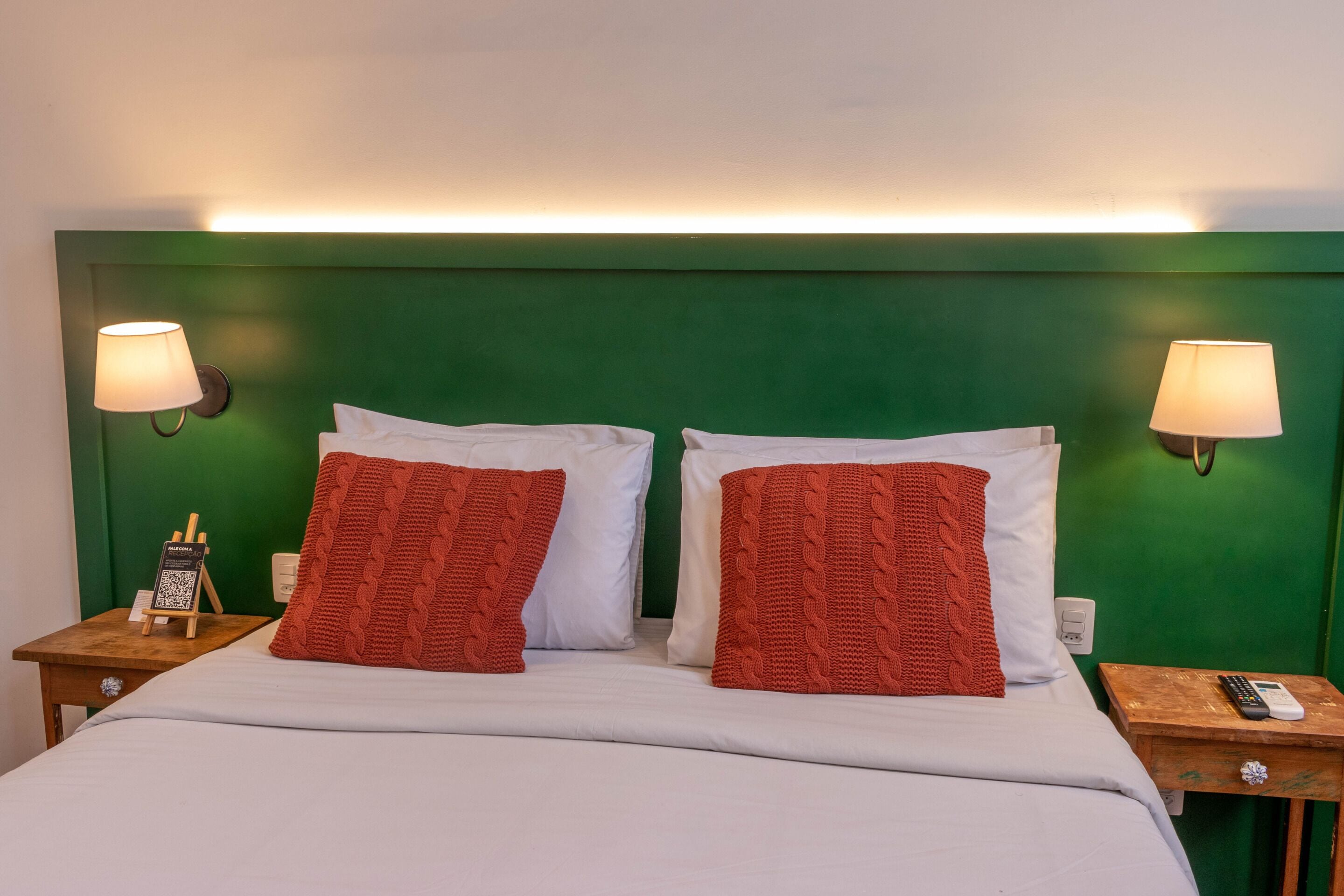 Une chambre d’hôtel avec une tête de lit verte, deux oreillers rouges, du linge de lit blanc, des lampes de chevet, une table de chevet avec un téléphone et une petite table avec une télécommande.