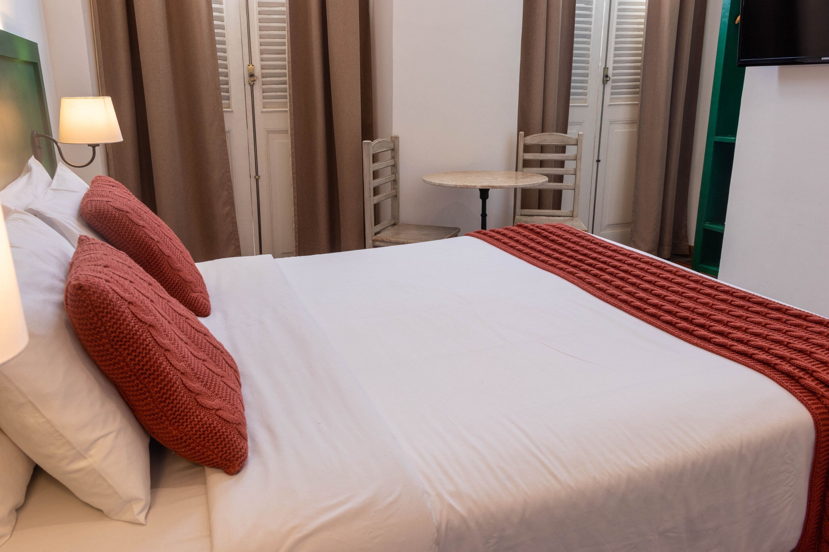 A neatly made bed with a white and red blanket, a small table with a chair, and a lamp on a bedside table.