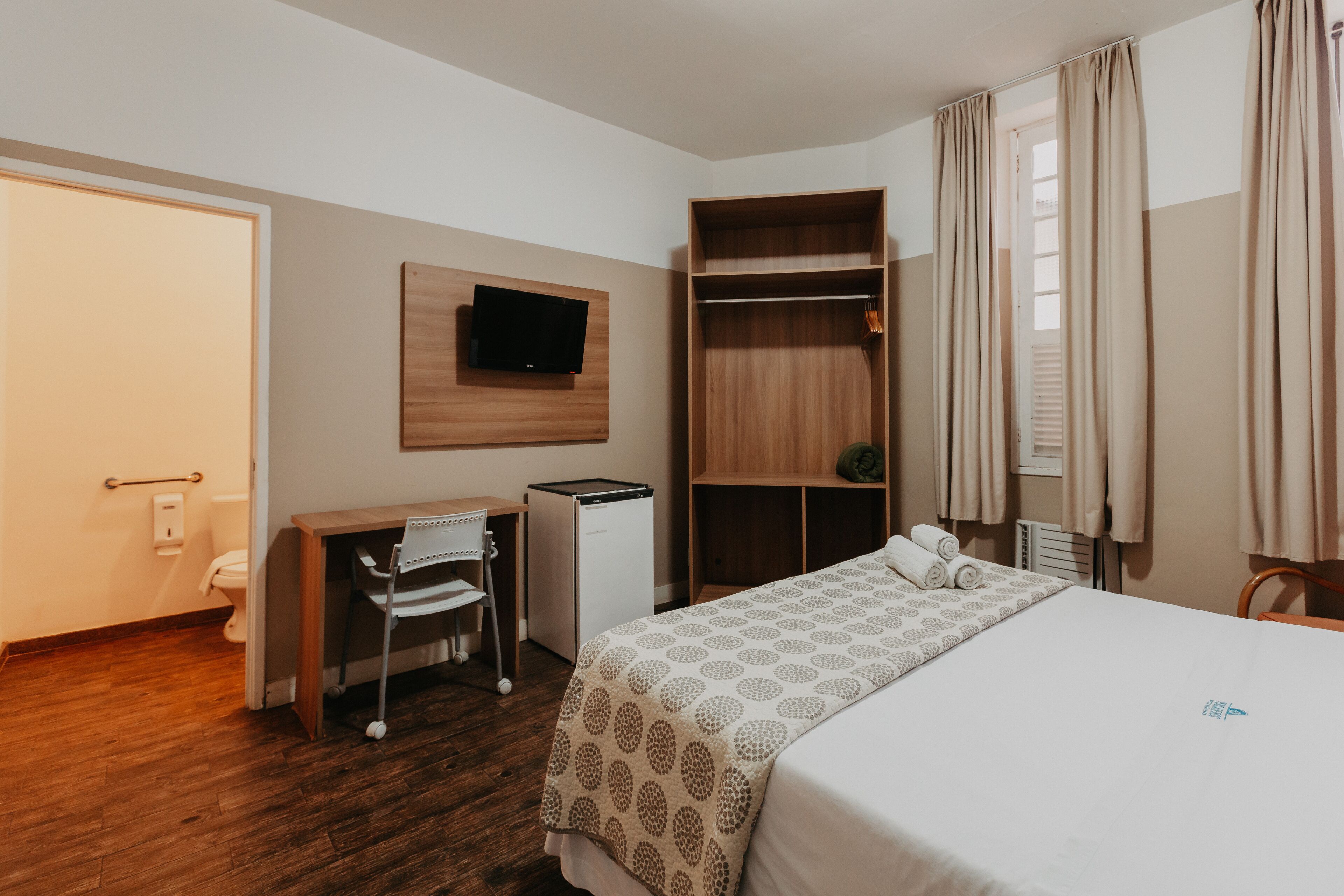 Habitación de hotel con cama, escritorio con silla, televisor montado en la pared, armario y ventana con cortinas.