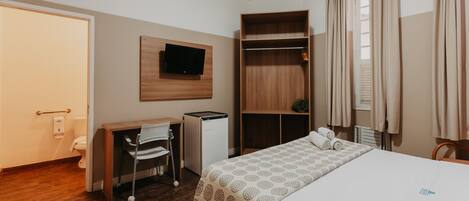 Habitación de hotel con cama, escritorio con silla, televisor montado en la pared, armario y ventana con cortinas.