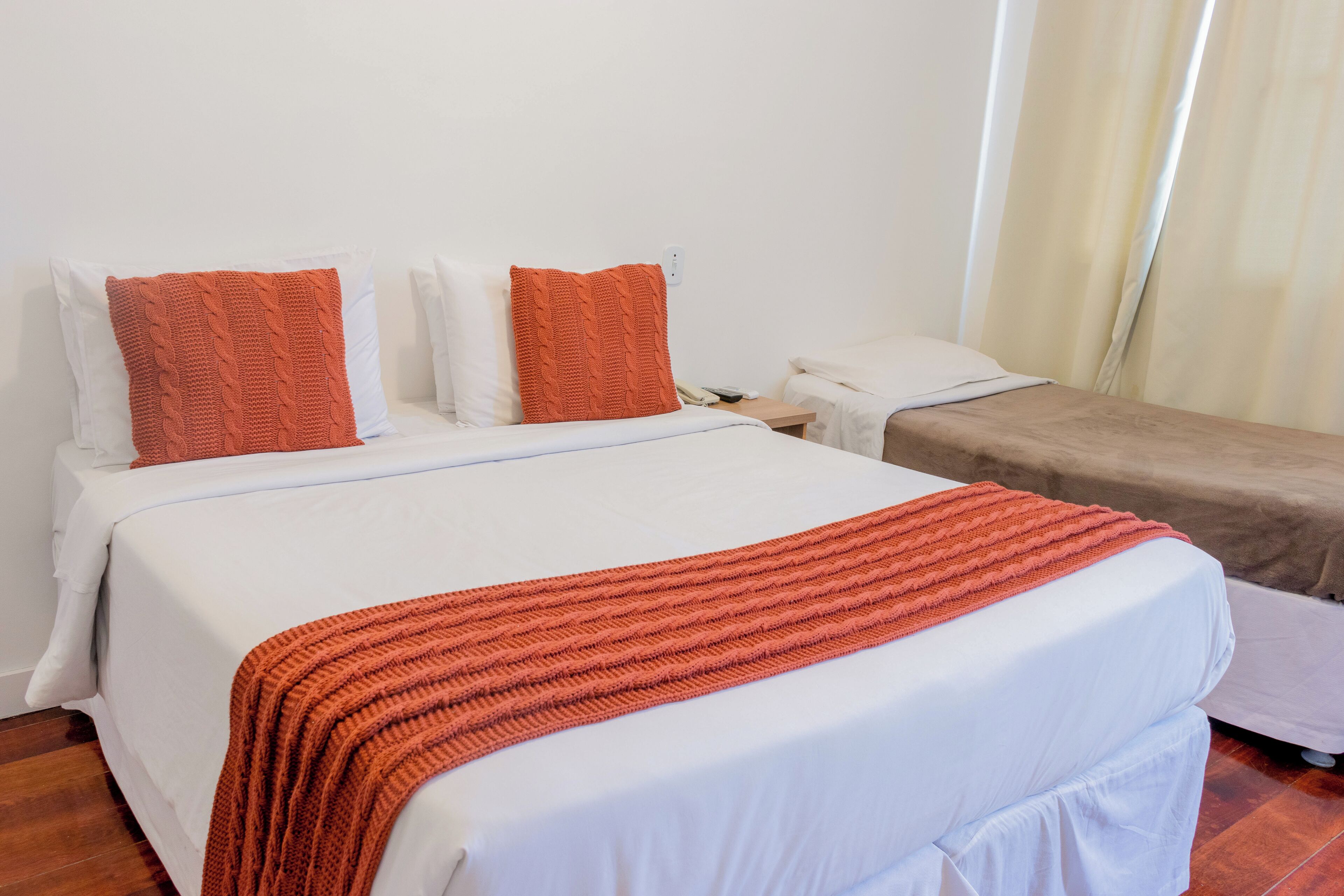 Ein Hotelzimmer mit einem großen Bett, einem kleineren Bett, weißer Bettwäsche und orangenen Zierkissen.