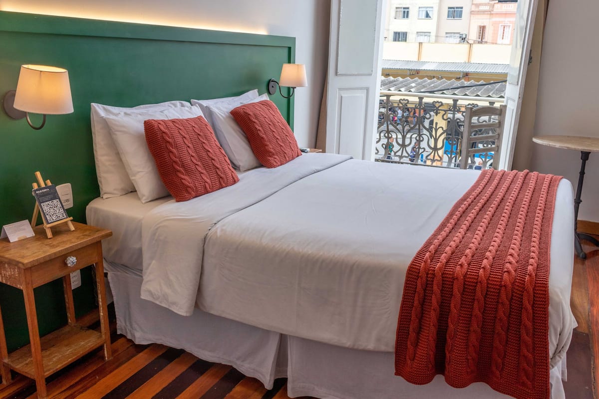 A neatly made bed with white linens and a red knitted throw, a wooden nightstand with a lamp, and a view of a balcony with metal railings.