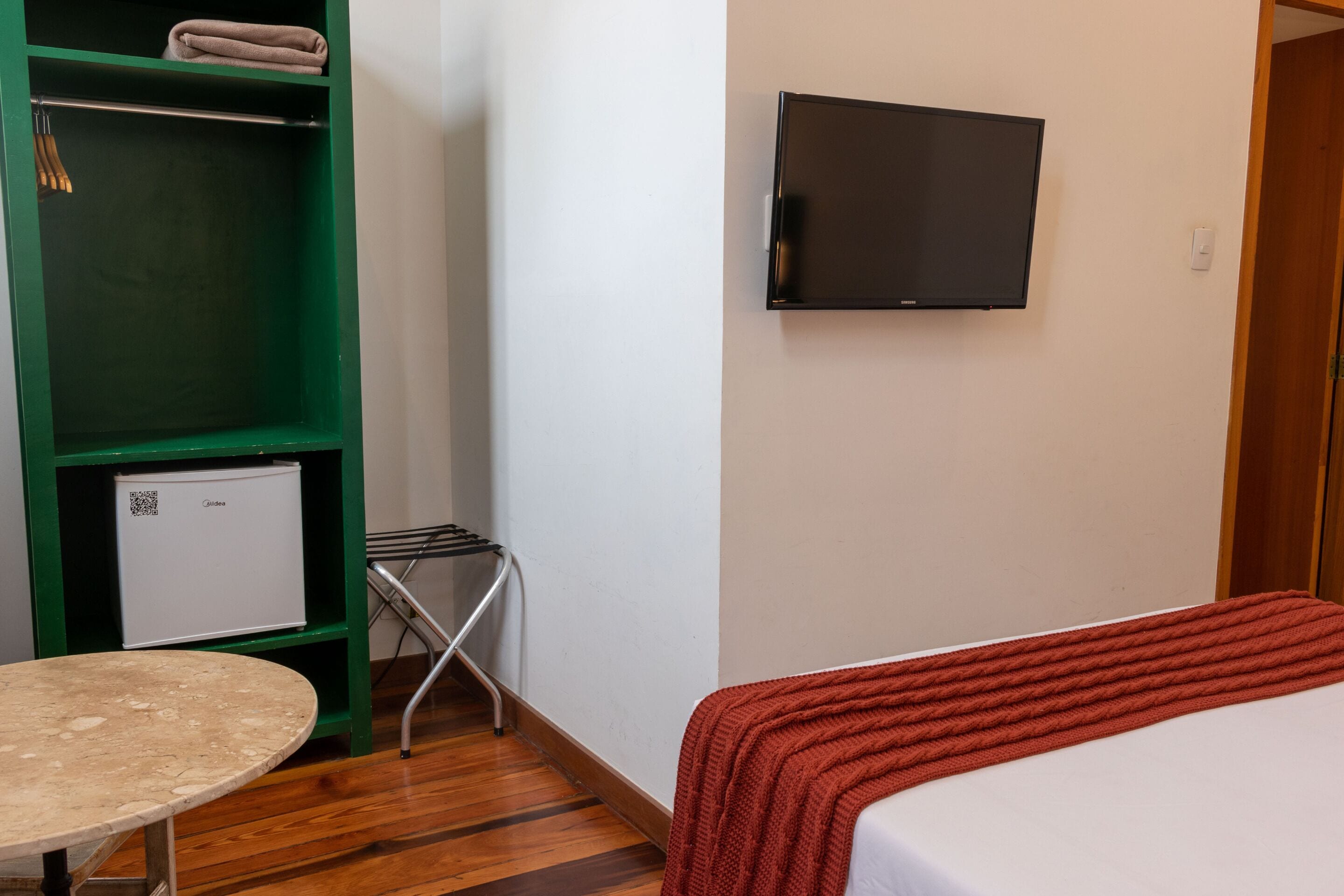 A hotel room with a bed, a television mounted on the wall, a green wardrobe, a small table, and a refrigerator.