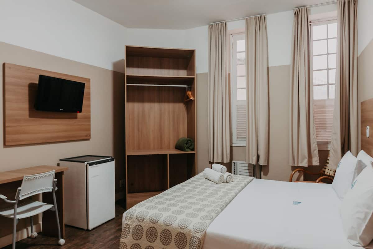 A hotel room with a bed, a desk, a chair, a television mounted on the wall, and a wardrobe.