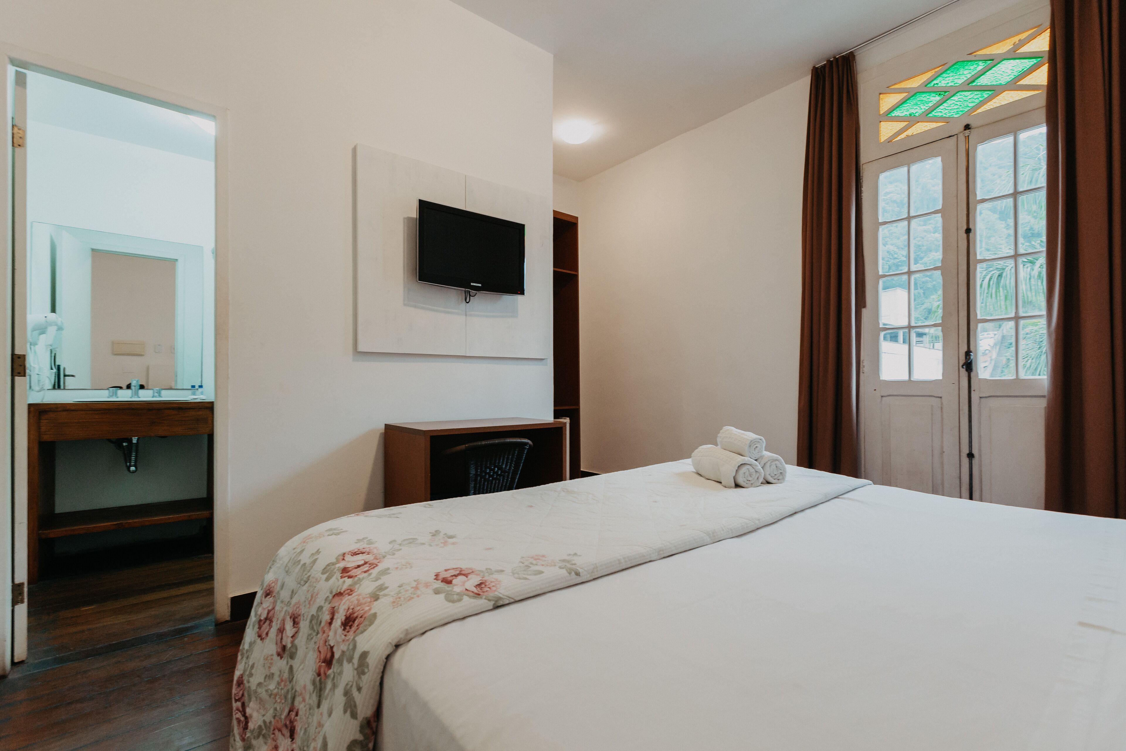 Quarto de hotel com cama, televisão na parede, escrivaninha, espelho e janela com cortinas.
