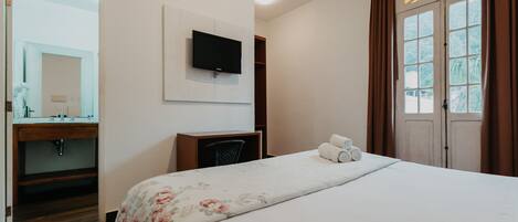Habitación de hotel con cama, televisor montado en la pared, escritorio, espejo y ventana con cortinas.