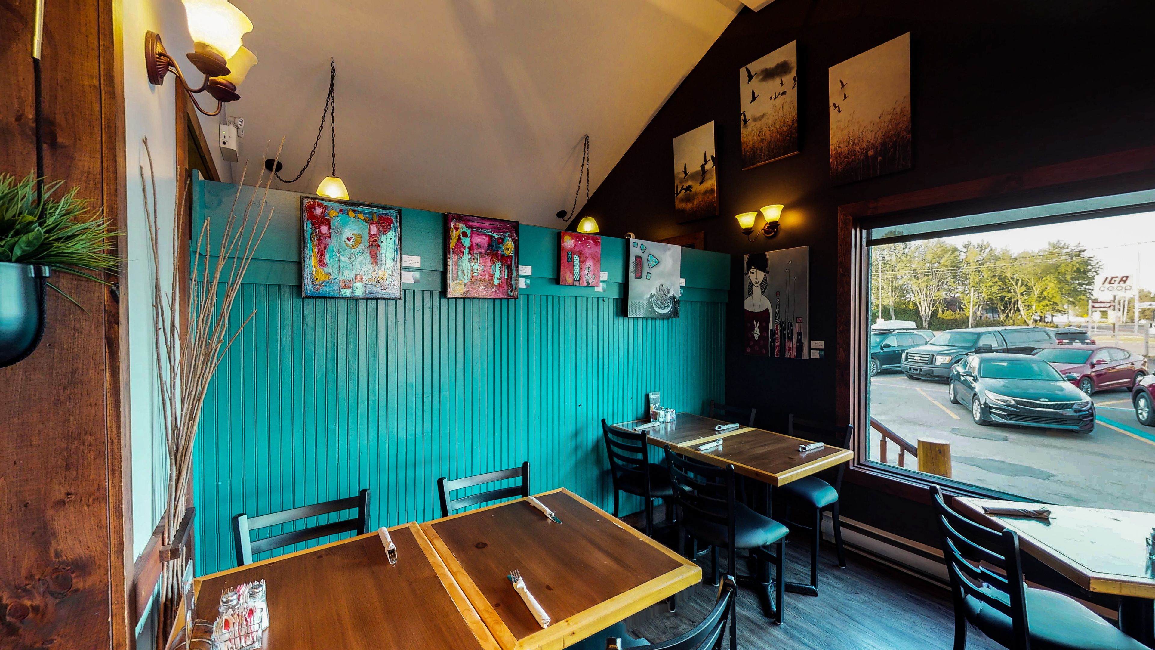 Interior de restaurante con paredes verde azulado, mesas y sillas de madera. Hay obras de arte enmarcadas en las paredes y se ve coches aparcados afuera.