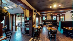 Interior de un restaurante con mesas y sillas de madera, una zona de bar y grandes ventanales con vistas.
