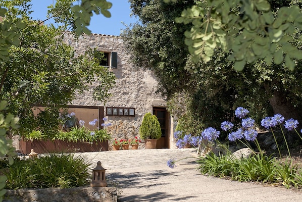 Un edificio rústico de piedra con puerta de madera, rodeado de abundante vegetación y plantas en flor.