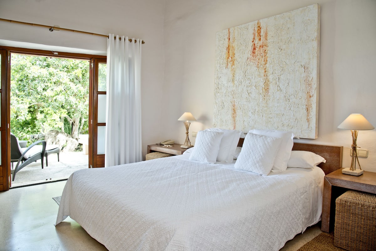 Un dormitorio amplio con una cama grande, dos mesitas de noche con lámparas, vistas a la vegetación a través de una puerta corredera y un cuadro abstracto de gran tamaño en la pared.