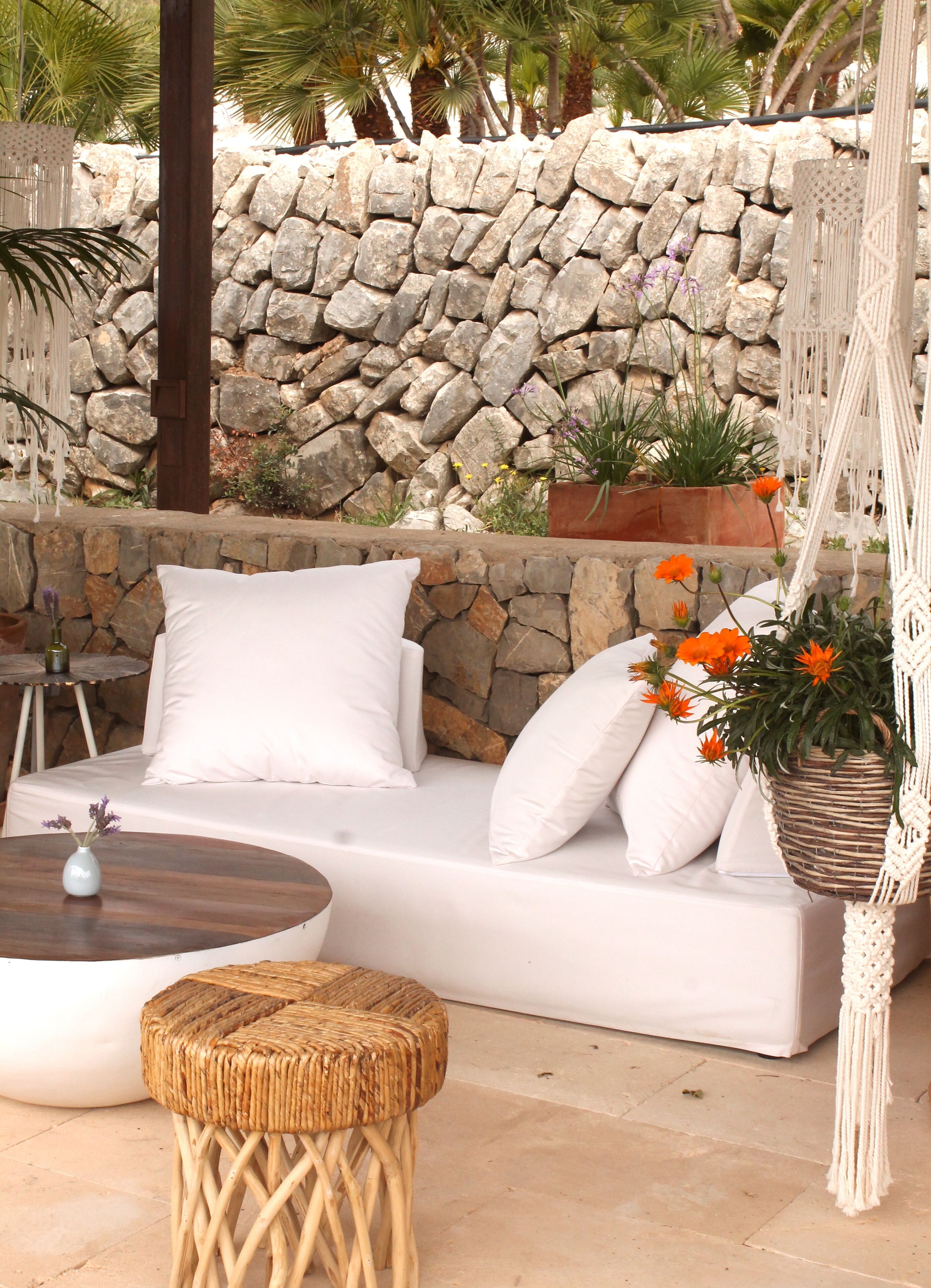 Un espacio exterior con mobiliario blanco, un muro de piedra al fondo, un taburete tejido y una decoración de macramé colgante.