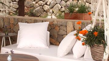 Un espacio exterior con mobiliario blanco, un muro de piedra al fondo, un taburete tejido y una decoración de macramé colgante.