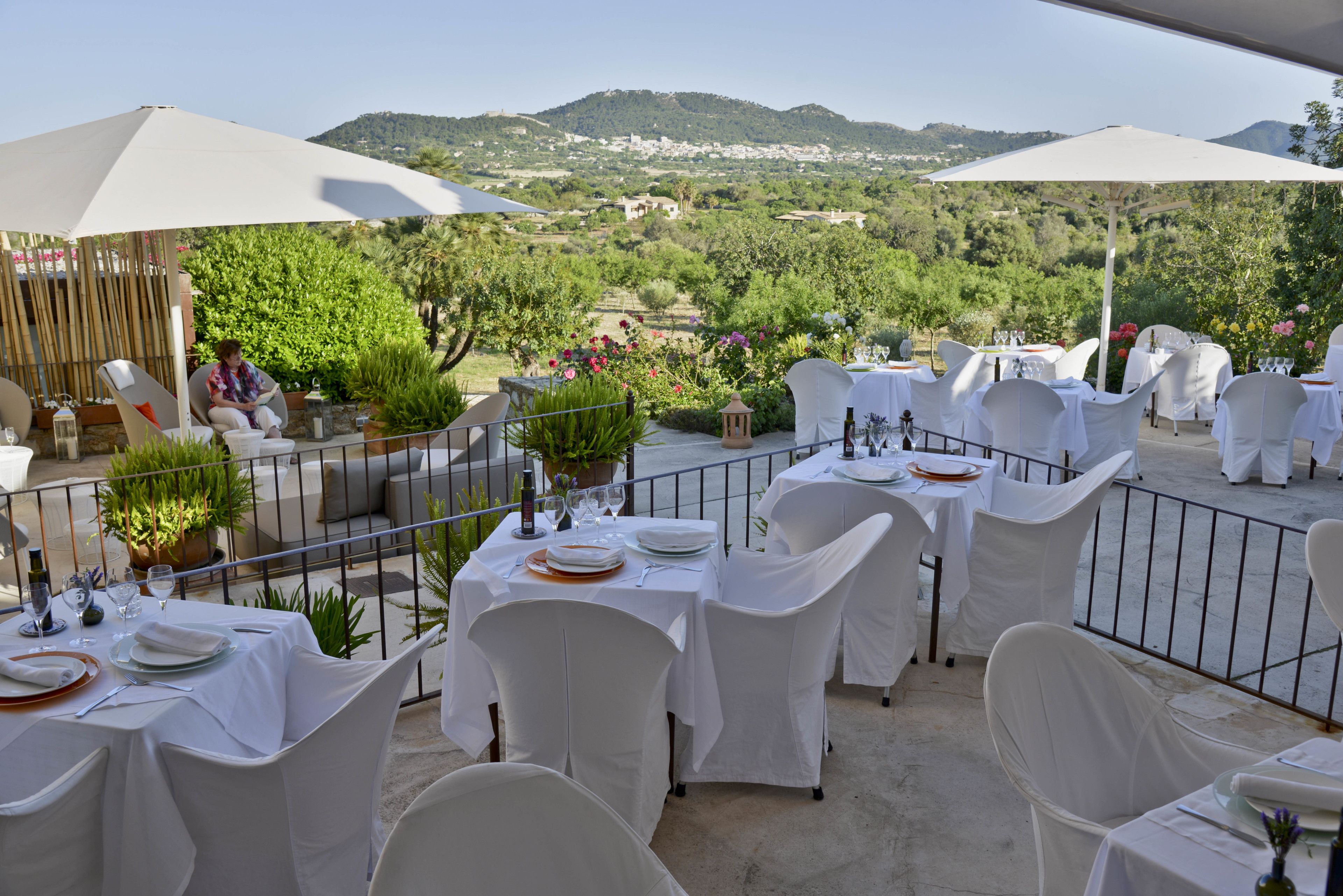 Un área de comedor al aire libre con mesas dispuestas para una comida, rodeada de sillas blancas y cubiertas con manteles blancos.