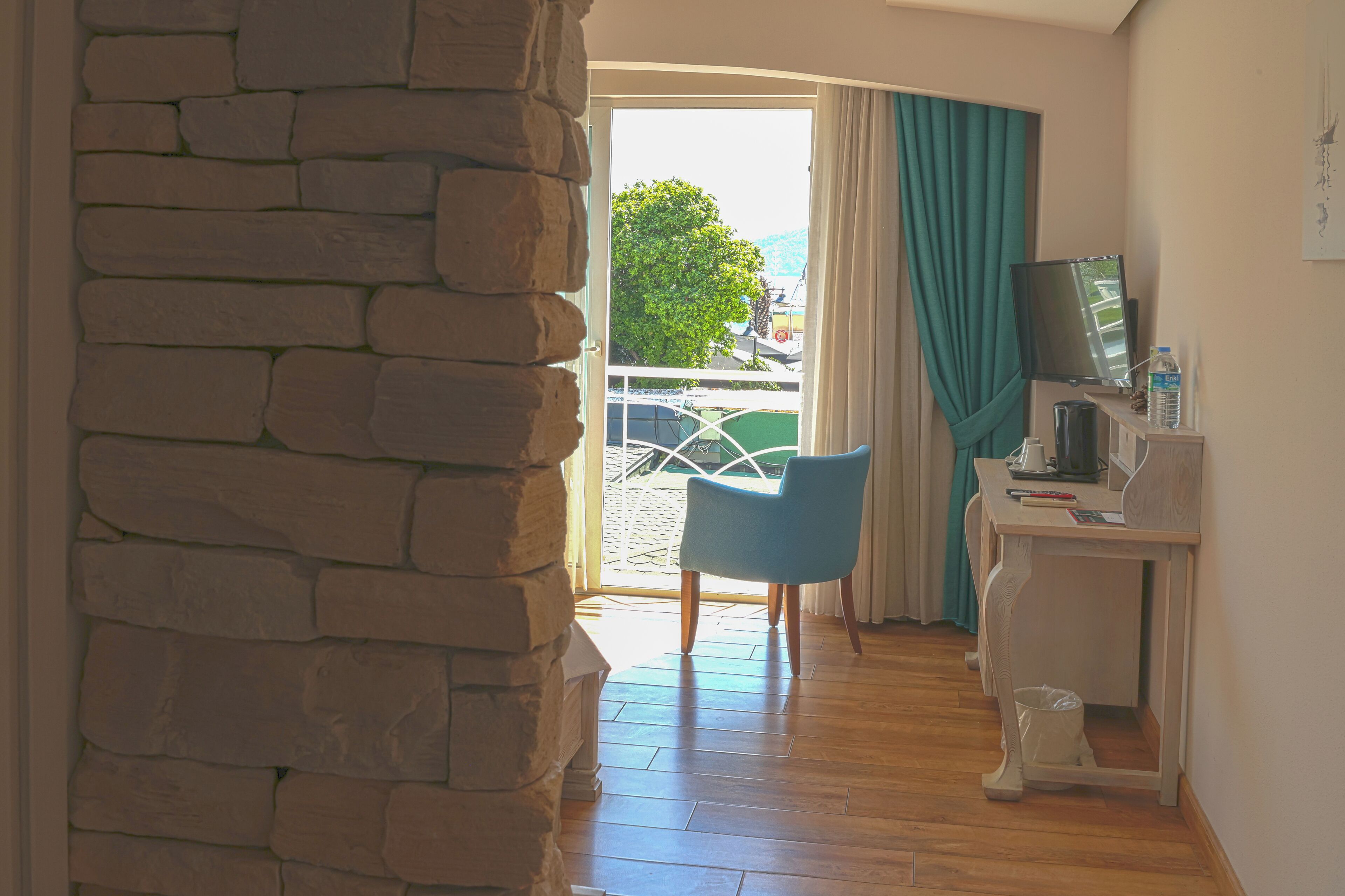 Una habitación con pared de piedra, una silla azul, un escritorio con televisor y vista a un árbol y coches aparcados a través de una puerta de vidrio.
