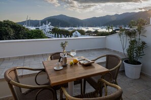 A balcony with wicker chairs, a wooden table, a potted plant, and a view of boats and mountains. - Dedeminn Marina Hotel (Fethiye)