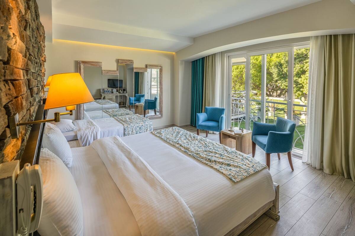 A hotel room with two beds, a sitting area with chairs, a TV, and a balcony with curtains.
