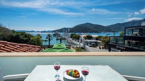 Una mesa con un plato de macarons de colores y dos copas de vino tinto, con vistas a la costa, barcos y montañas al fondo.