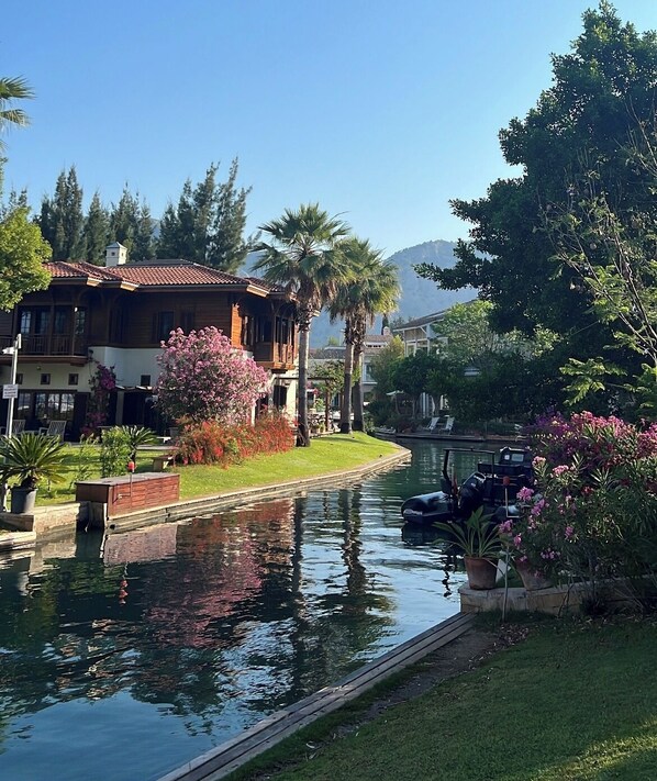 A canal with a wooden house on the left, surrounded by greenery and flowers.
