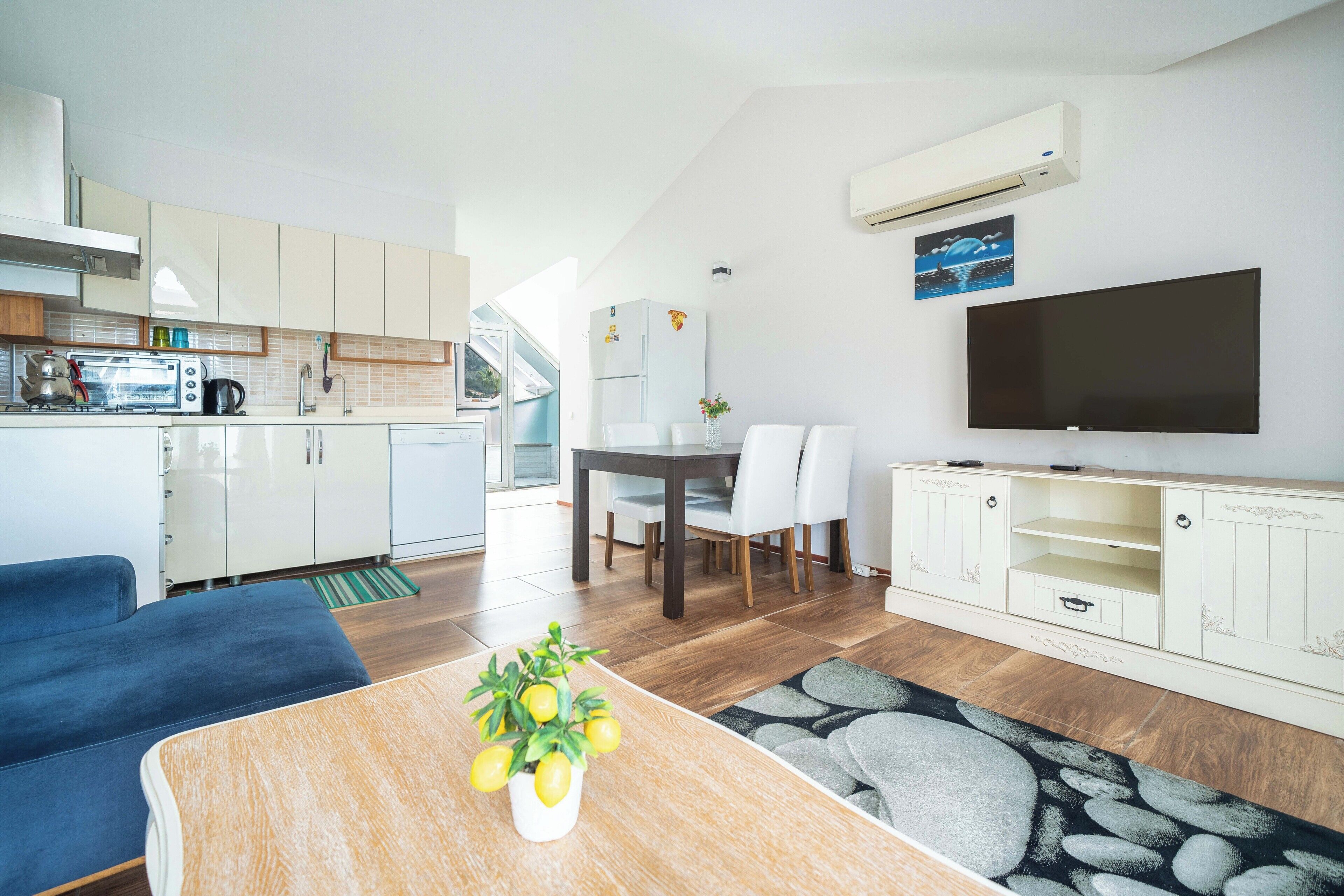 Una cocina moderna con armarios blancos, un comedor con mesa y sillas, un televisor de pantalla plana y una sala de estar con sofá y mesa de centro.