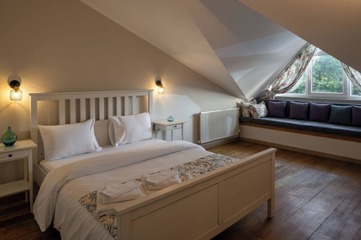 A bedroom with a sloped ceiling, a bed with white bedding, a nightstand, a small table, and a window with a view of greenery.