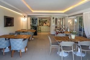 A modern dining area with wooden tables and chairs, a kitchen in the background, and large windows with curtains.