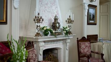 A room with a fireplace, ornate candelabras, and floral arrangements. There are two upholstered chairs and a table with a white tablecloth.